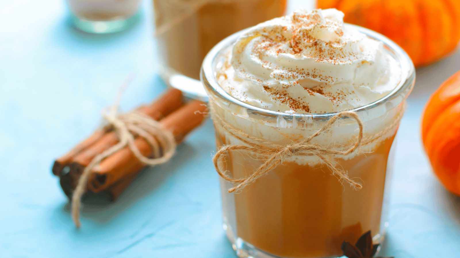 A glass of pumpkin spice latte, reminiscent of the warmth found in superfood smoothies, is topped with whipped cream and sprinkled with cinnamon. Two cinnamon sticks tied with twine sit next to the glass, while small orange pumpkins add a seasonal touch on a light blue surface.