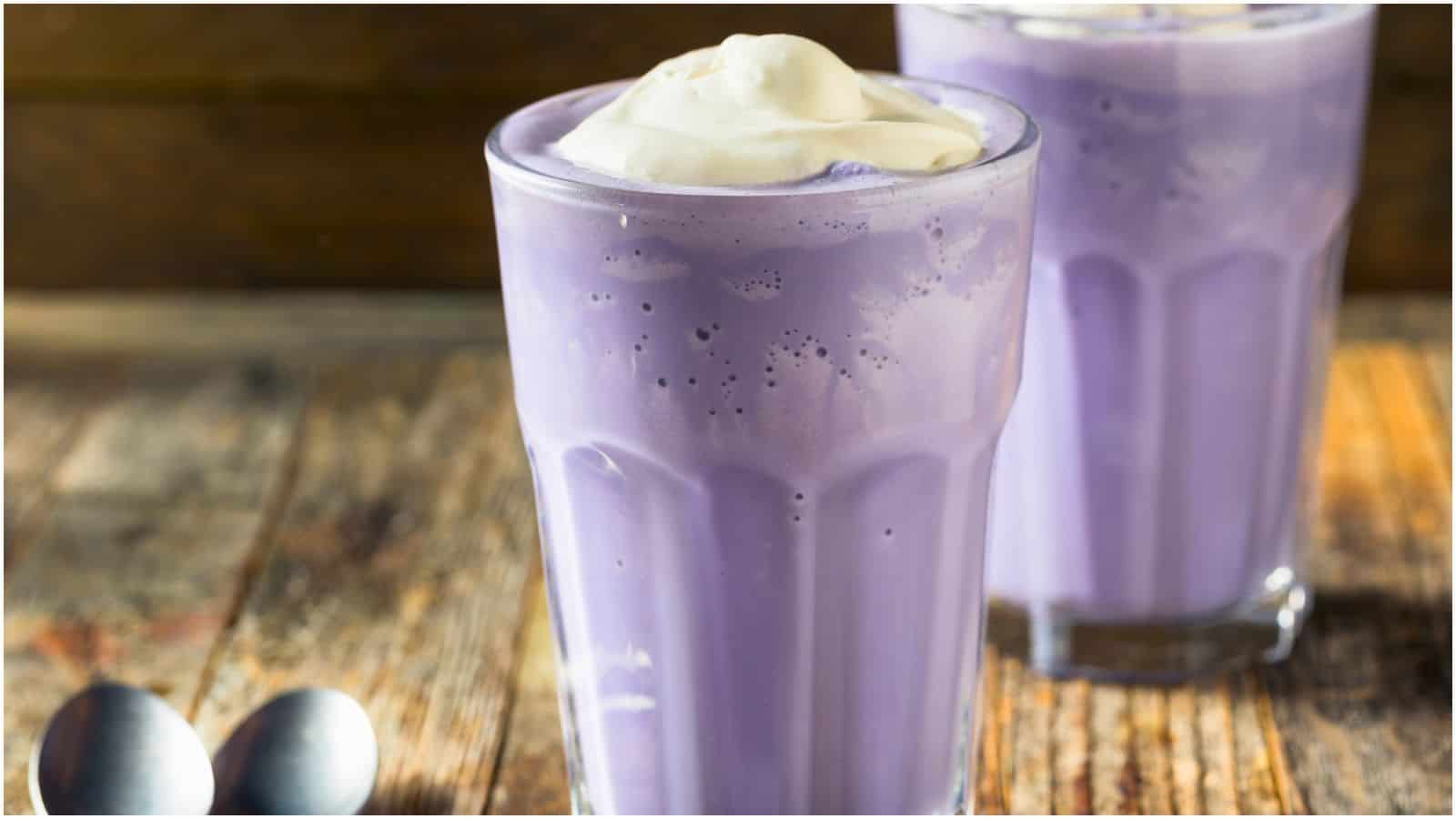 Two glasses of purple ube milkshakes are on a wooden table. Each milkshake is topped with a dollop of whipped cream. Two spoons are placed next to the glasses. The background is rustic wood.