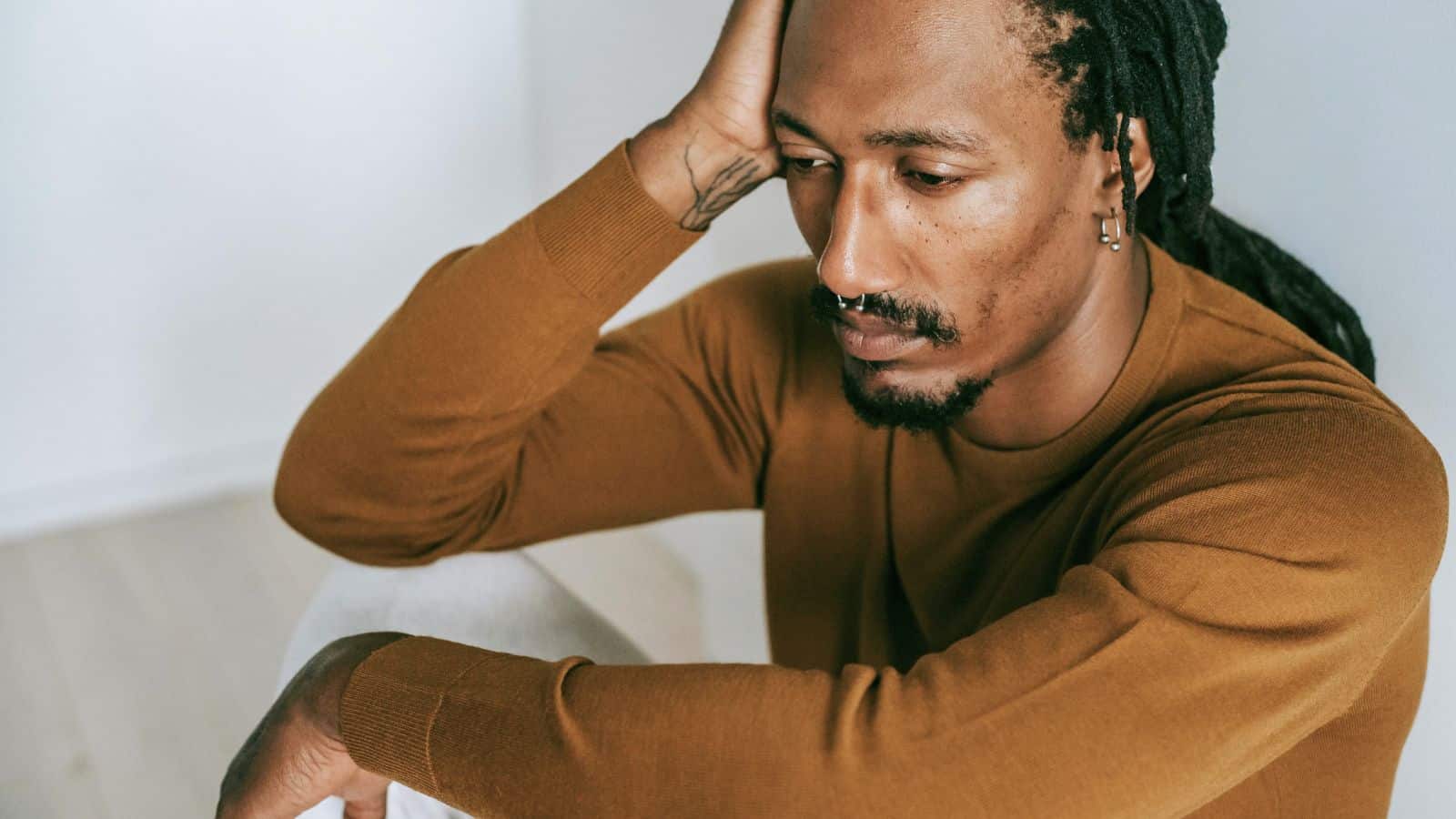 A person with long braided hair, a nose ring, and earrings is sitting on the floor, leaning their head on their left hand. They are wearing a long-sleeve brown shirt and light-colored pants, looking pensive.