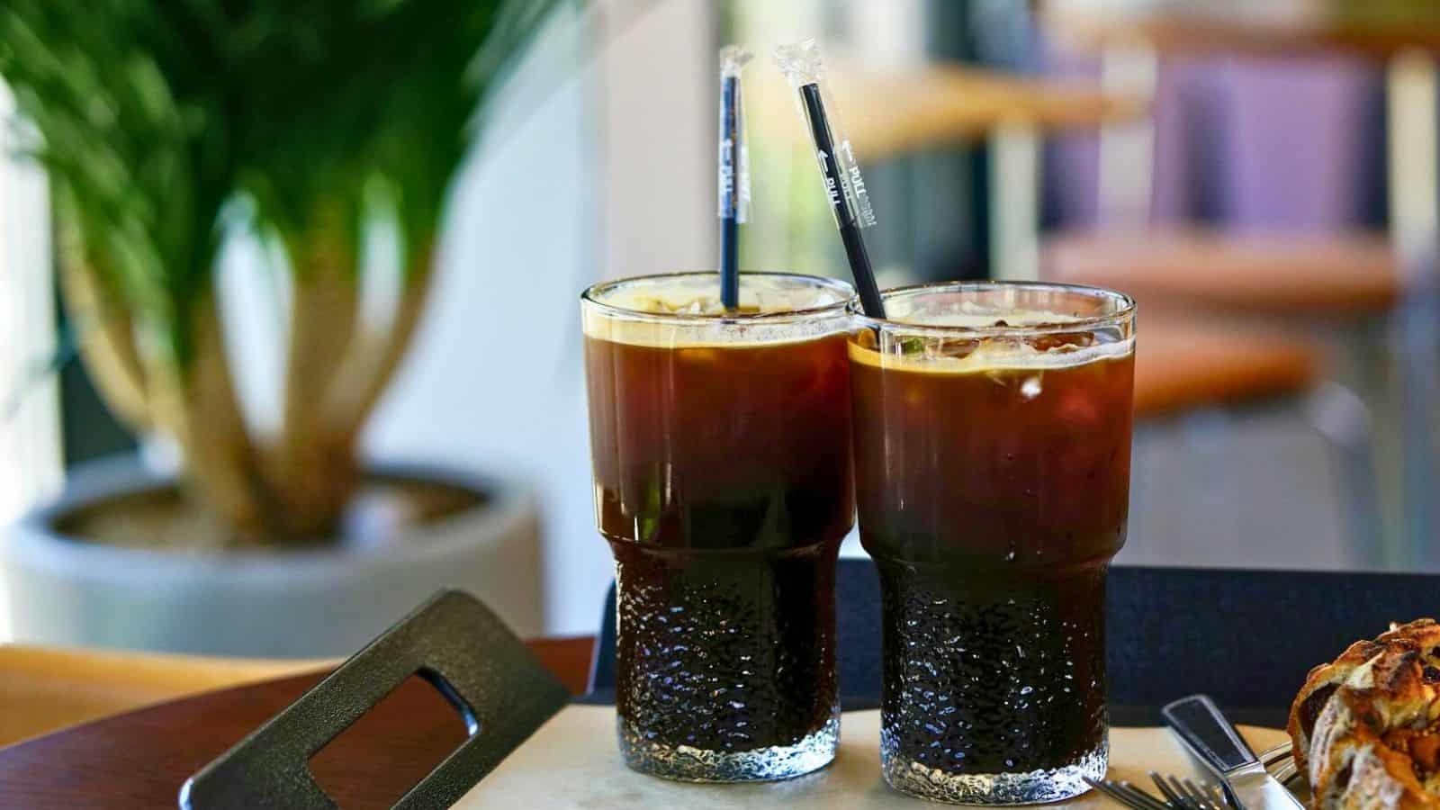 Two glasses of iced tea with black straws sit on a table. The glasses boast a textured design and condensation on the outside. In the background, there's a blurred view of indoor plants and chairs.
