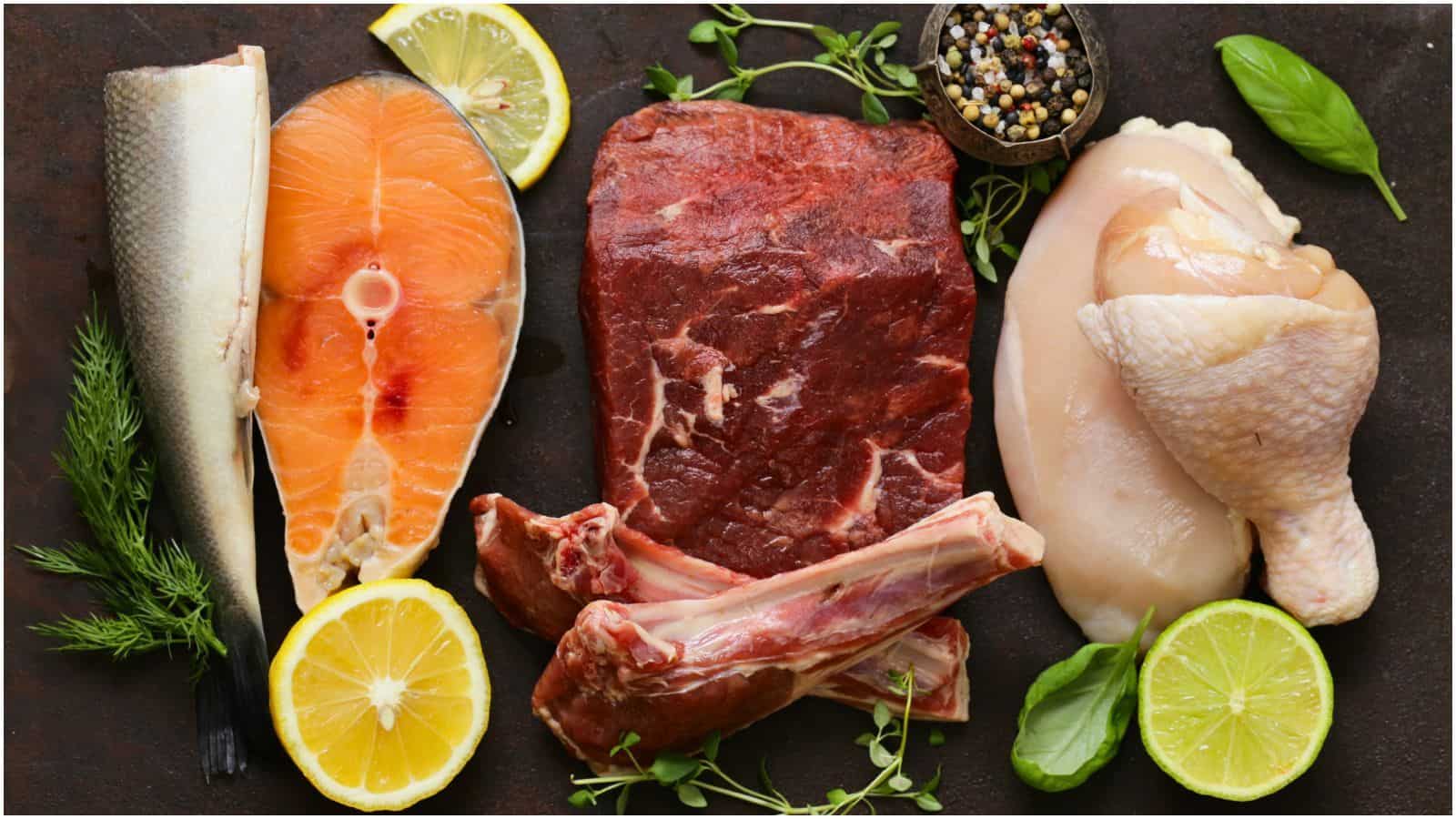 An assortment of raw meats and fish displayed on a dark surface. It includes a salmon fillet, a steak, chicken breast, and a piece of chicken leg. Herbs, lemon slices, and a small bowl of mixed peppercorns are arranged around the meats.