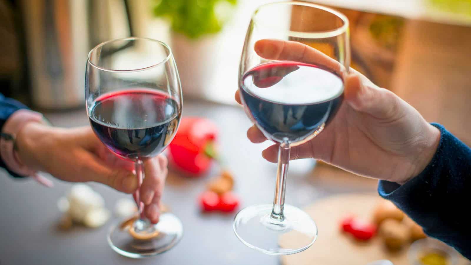 Two people clinking glasses of red wine over a table with a blurred background of various items including garlic, a red pepper, and greenery.