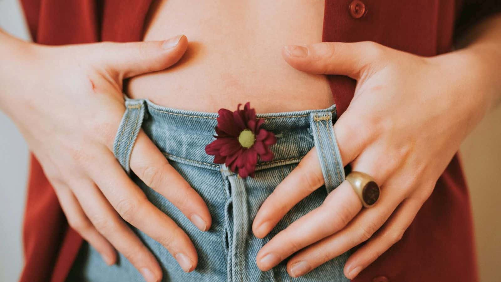 Close-up of a person with a flower in the waistband of their blue jeans. The individual is wearing a red shirt open at the midsection, with both hands resting on their jeans. 