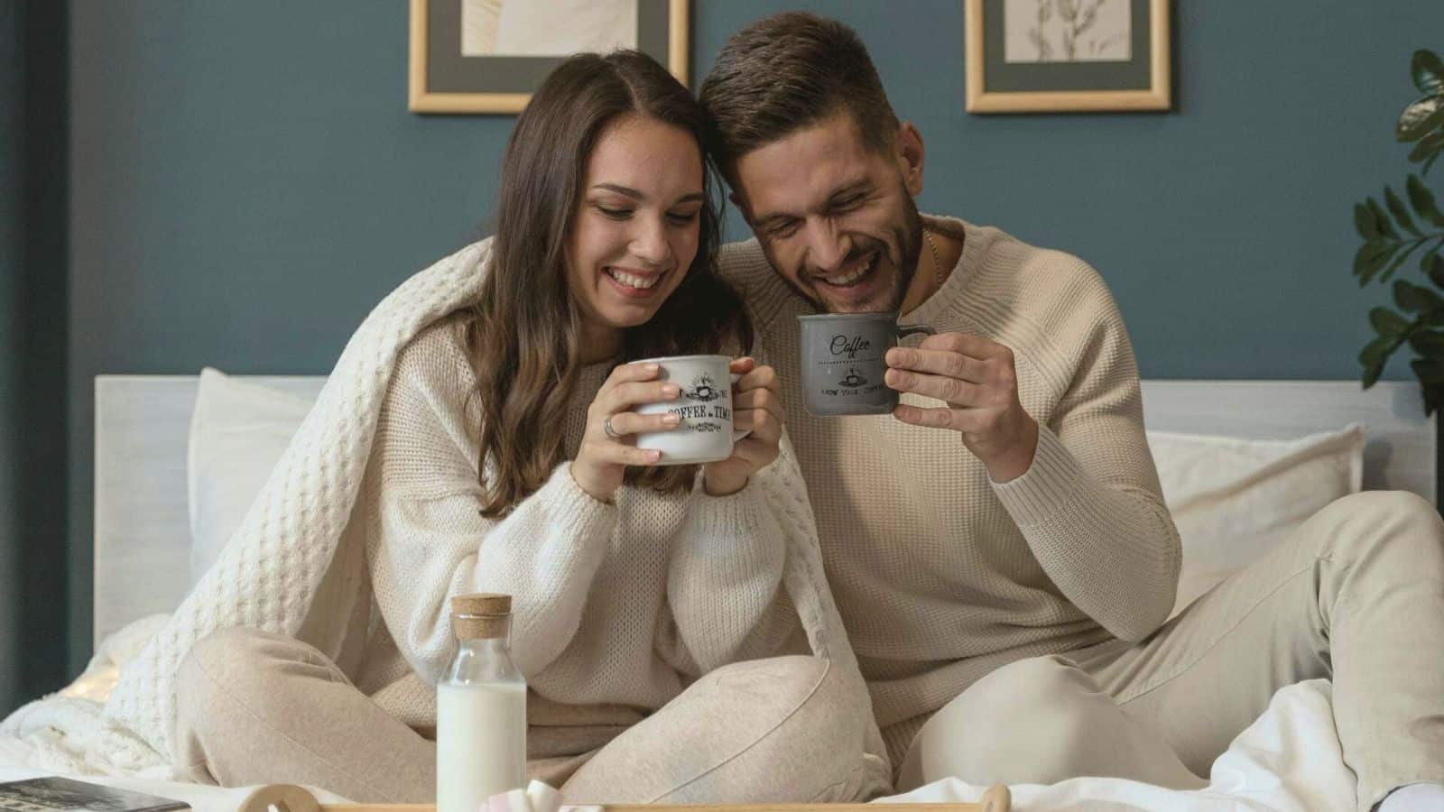 A man and woman, both dressed in beige casual clothing, sit close together on a bed, smiling and holding mugs. There is a bottle of milk and a candle on the bedside table. 