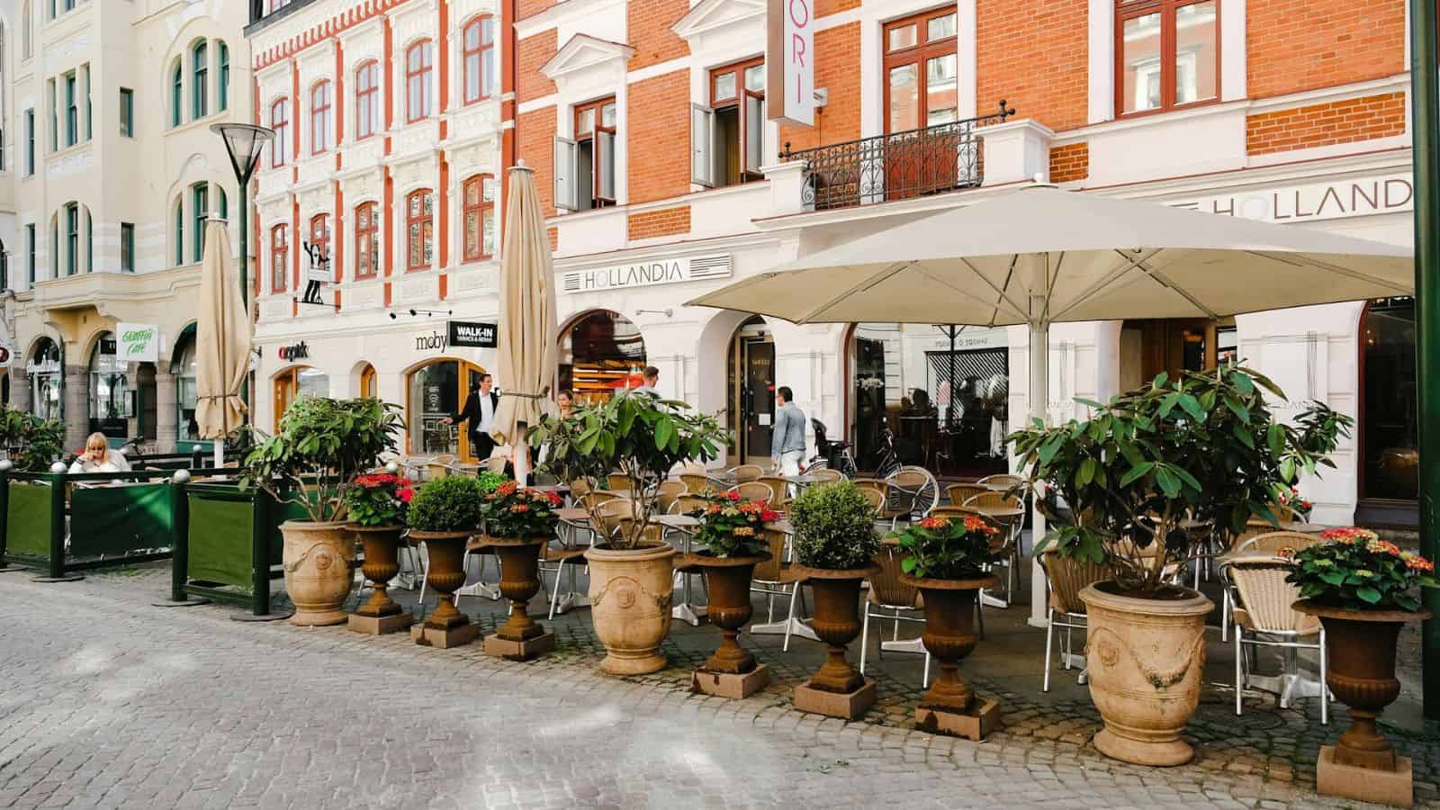 Outdoor cafe with large umbrellas and tables surrounded by potted plants on a cobblestone street. The scene is set against a backdrop of colorful, multi-story buildings with storefronts and windows. A few people are seated and walking nearby.
