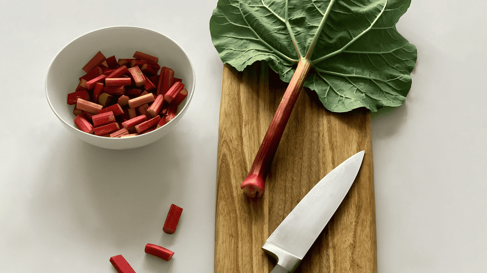 A large rhubarb leaf and a cutting board with a kitchen knife are on display. A bowl brims with chopped rhubarb, suggesting the transformation of these unusual plants for winemaking, alongside some additional segments on the board.