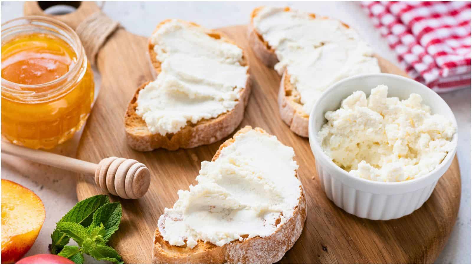Image of a wooden board with slices of bread spread with ricotta cheese. A white ramekin filled with more ricotta, a jar of honey, and a honey dipper are placed beside the bread. Fresh mint leaves and a peach slice are also visible on the board.