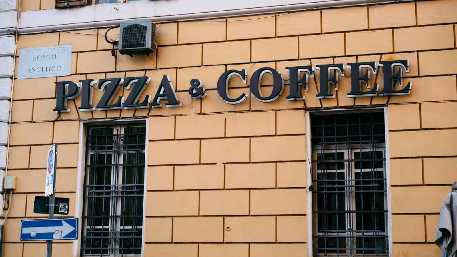 The image shows a building with a sign reading "Pizza & Coffee" above two barred windows. There's a street signpost in front and a small sign to the left of the windows. An air conditioning unit is mounted above the sign.