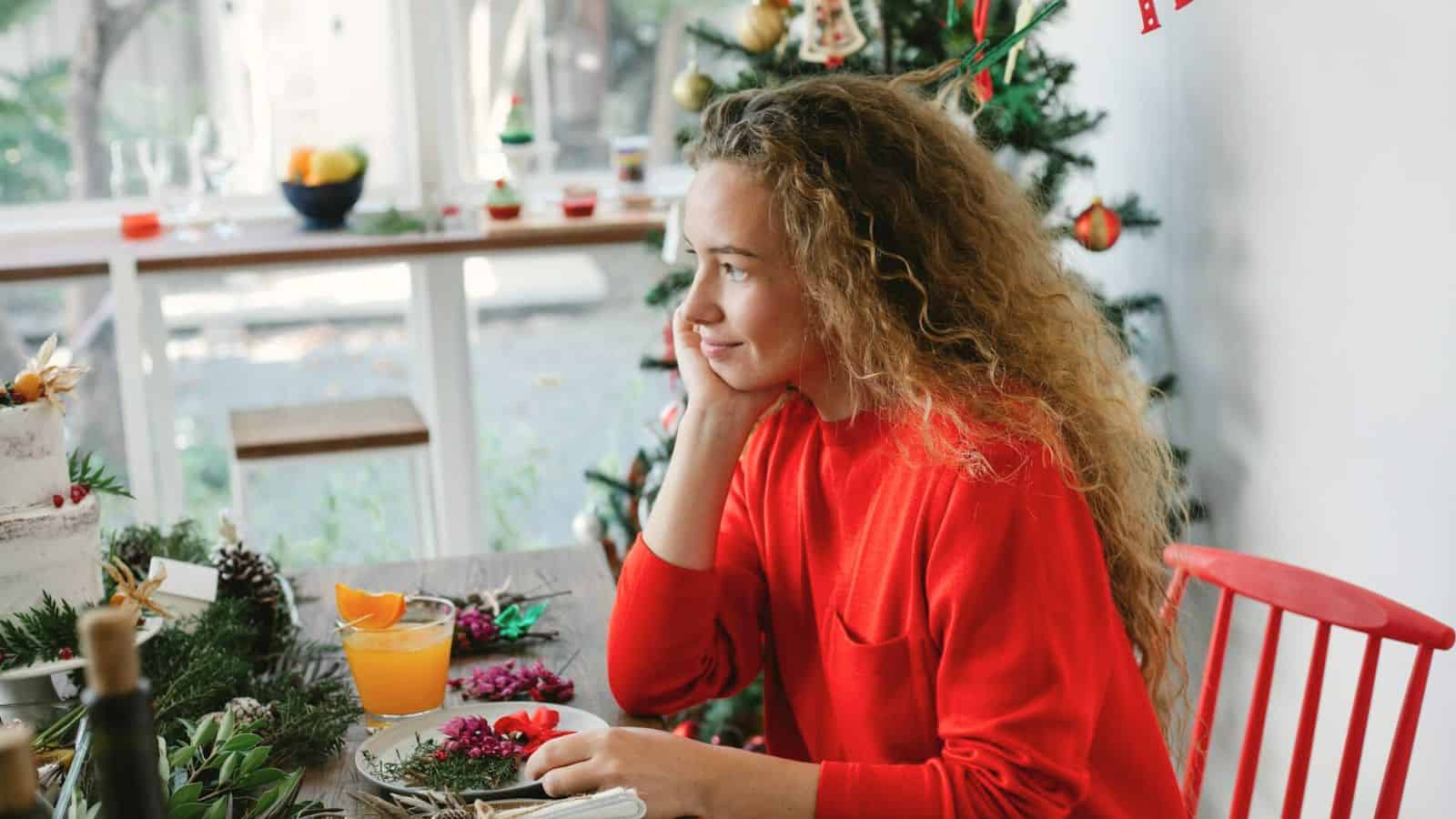 A woman with curly hair, wearing a red sweater, sits at a table by a decorated Christmas tree. She rests her chin on her hand, looking out of the window. The table has festive decorations,  a slice of cake, and a glass of orange juice.