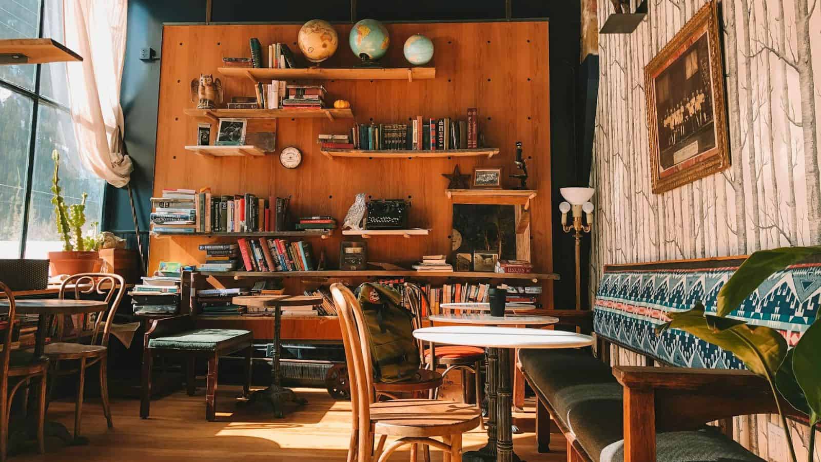 A cozy room with bookshelves filled with books and globes. There are wooden chairs and tables, including a white round table in the center. A patterned bench lines the right wall, with a framed picture above it. Sunlight streams in from a window.