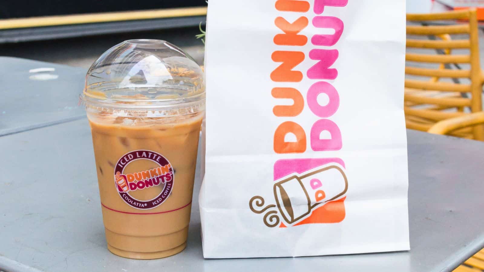 A Dunkin' Donuts iced latte in a plastic cup with a dome lid sits on a table next to a white Dunkin' Donuts bag featuring the company's logo and branding.