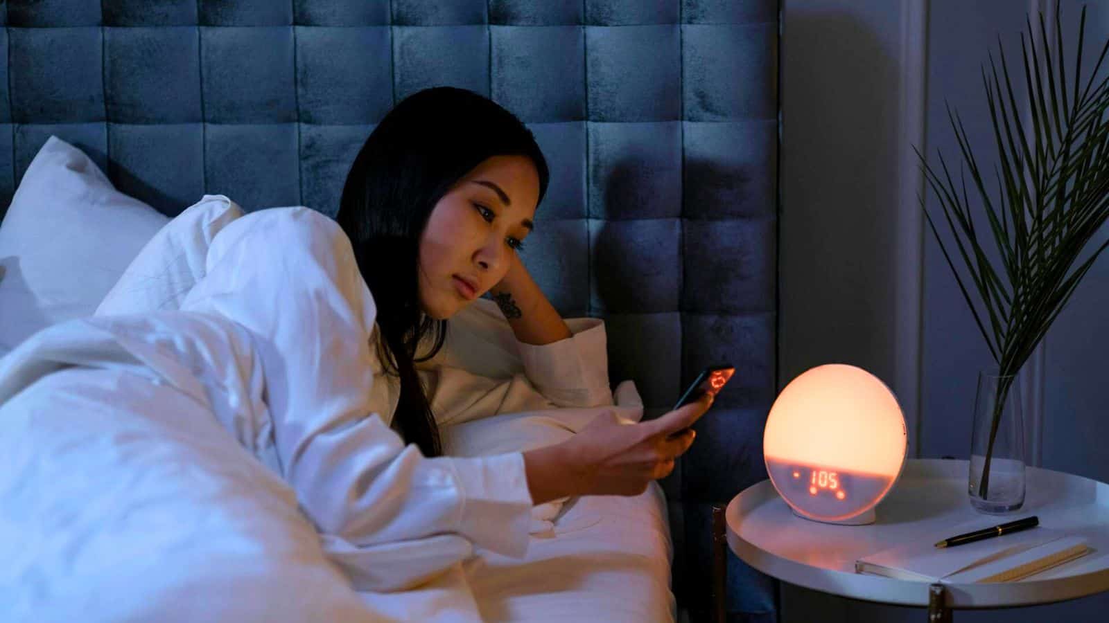 A person lies in bed with a tufted headboard, looking at a smartphone. A round digital clock displaying "1:05" and a leafy plant in a clear vase are on a small bedside table. The room is dimly lit.