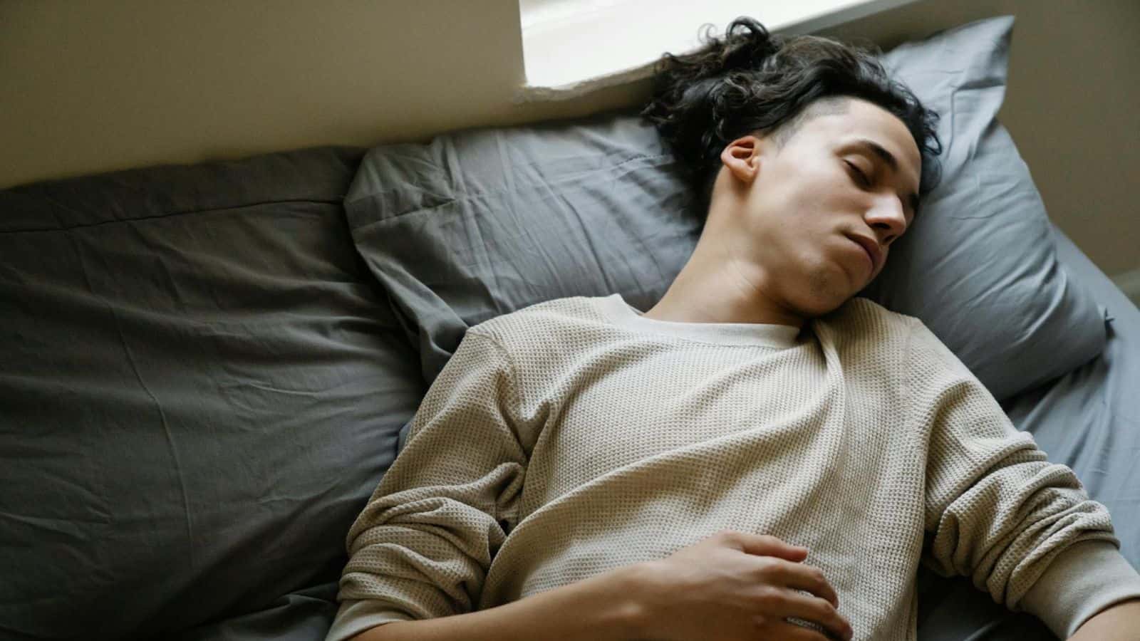A person with long hair lies asleep on a bed with gray sheets, resting their head on a pillow. They are wearing a long-sleeved beige shirt and are positioned towards the edge of the pillow, one arm resting on their torso.