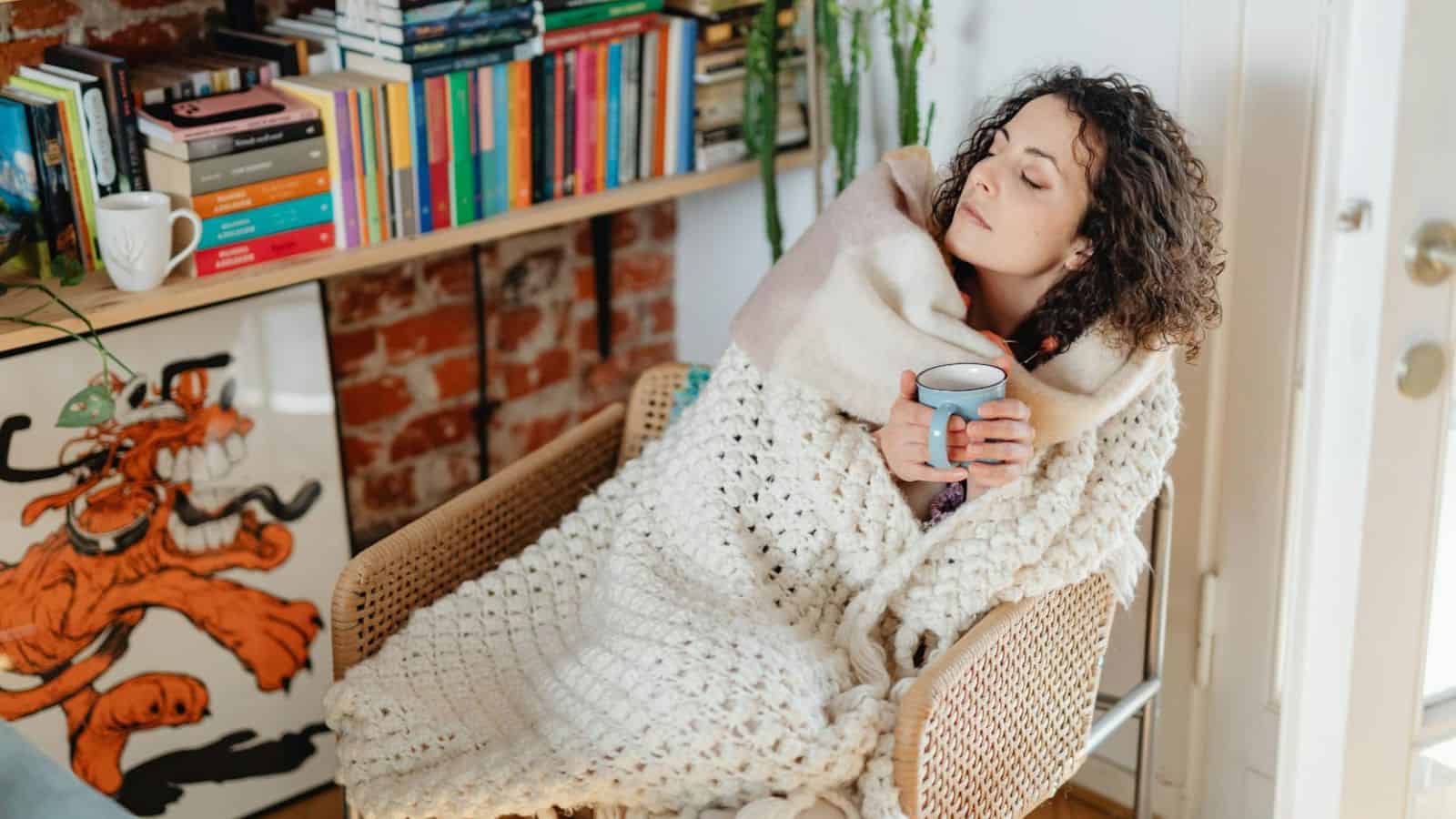 A person with curly hair sits in a wicker chair, wrapped in a thick blanket, holding a blue mug. Behind them is a bookshelf filled with colorful books and a framed cartoon image of a dog. Nearby, a green plant adds a touch of nature to the scene.