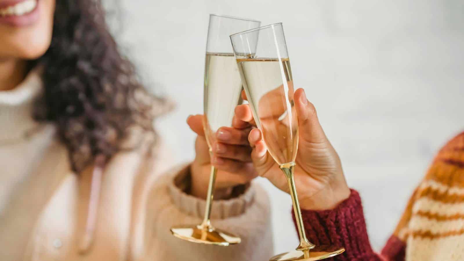 Two people clink champagne glasses in a toast, each dressed in cozy sweaters&mdash;one beige, the other maroon with a patterned design. Both glasses contain a clear, sparkling beverage.