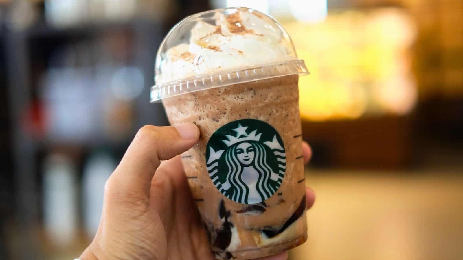 A hand holding a Starbucks iced beverage with whipped cream on top. The drink is in a clear plastic cup with a dome lid, displaying the Starbucks logo. The mocha or chocolate-flavored drink, possibly one of the Sta
