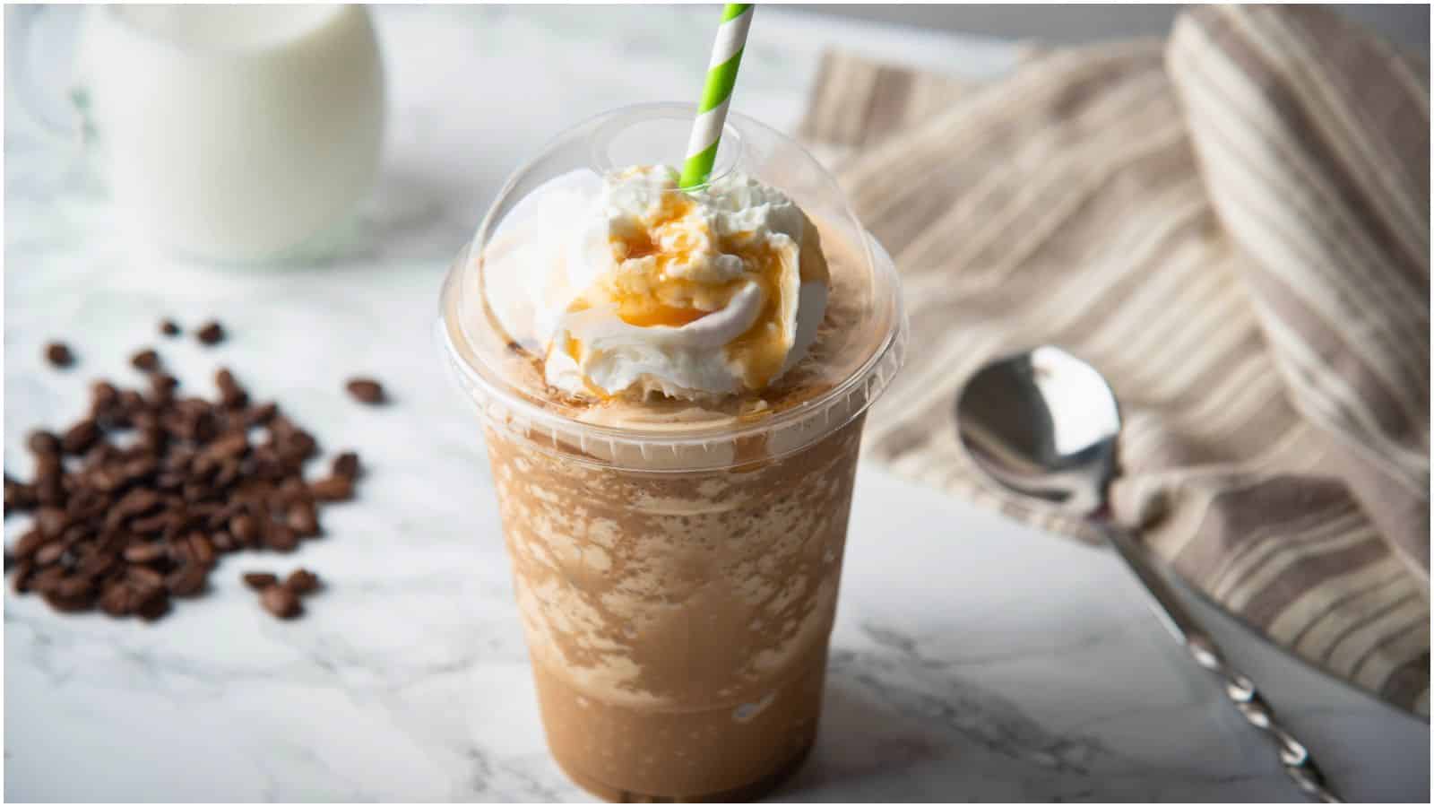 A close-up of a blended iced coffee drink topped with whipped cream and caramel drizzle in a transparent cup with a green and white striped straw. It is placed on a marble countertop next to a spoon, a striped cloth, and some scattered coffee beans.