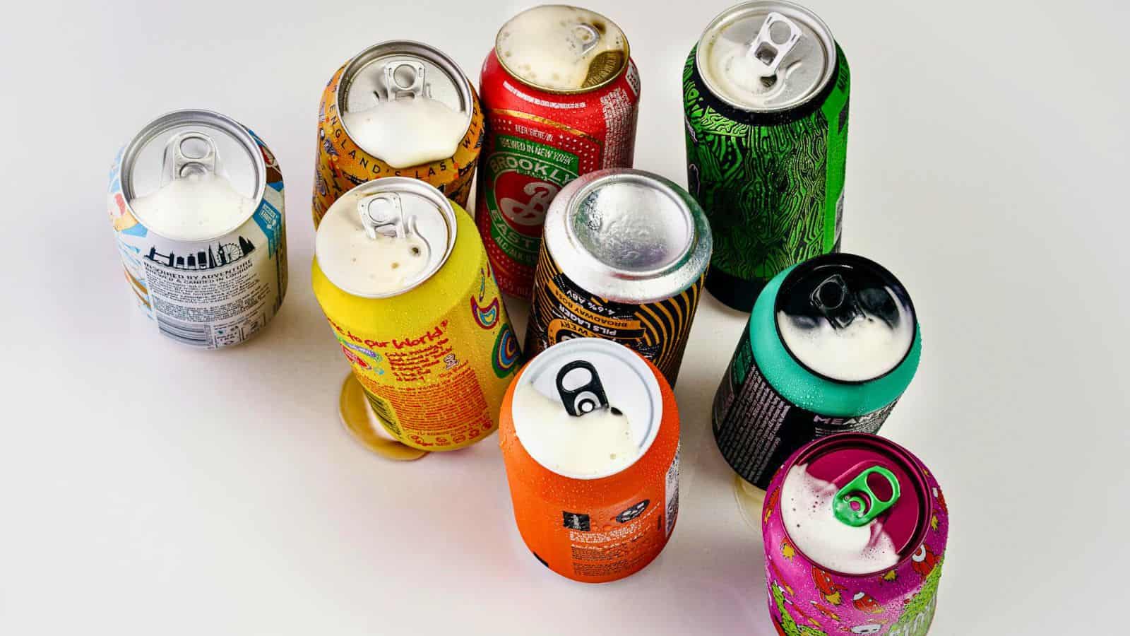 A collection of nine colorful opened cans containing various drinks arranged on a white surface. Each can has a distinct design and label, with some showing foam visible at the opening.