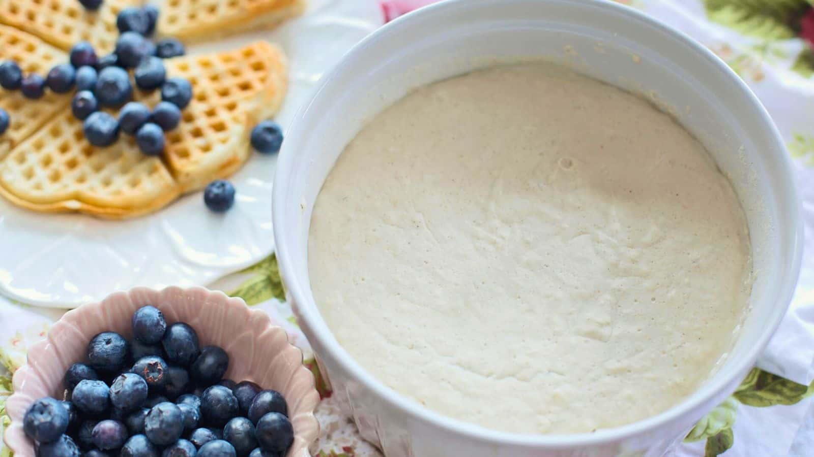 A bowl filled with sour cream is surrounded by a plate of heart-shaped waffles topped with blueberries and a small bowl of fresh blueberries. The setting is on a table with a floral tablecloth.