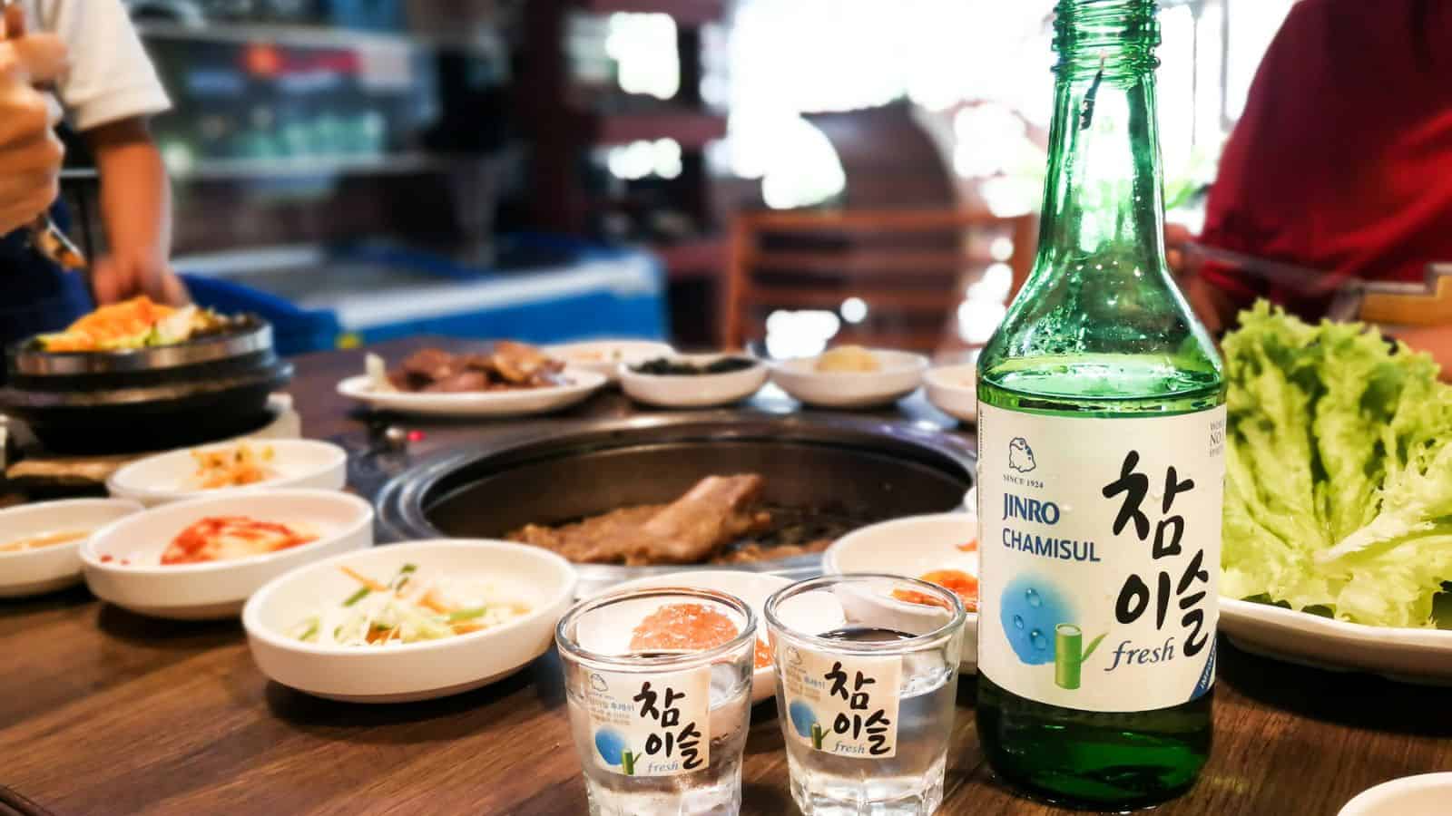 A tall bottle of soju with two shot glasses beside it. Behind them, there is a korean barbecue setting. 