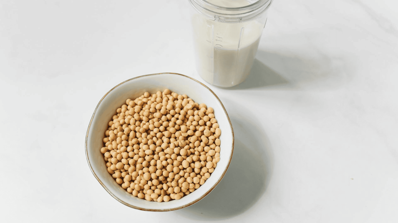 A white bowl filled with soybeans sits beside a clear glass of plant-based dairy milk substitute, soy milk, on a white surface.