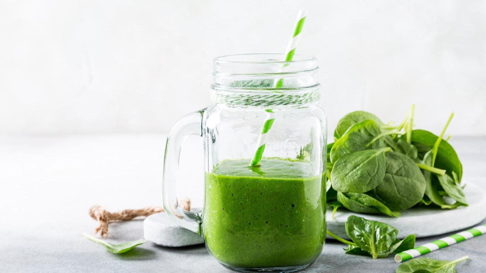 A mason jar filled with a green juice, equipped with a green and white striped straw. Fresh spinach leaves are placed beside the jar on a light gray surface with a neutral background.