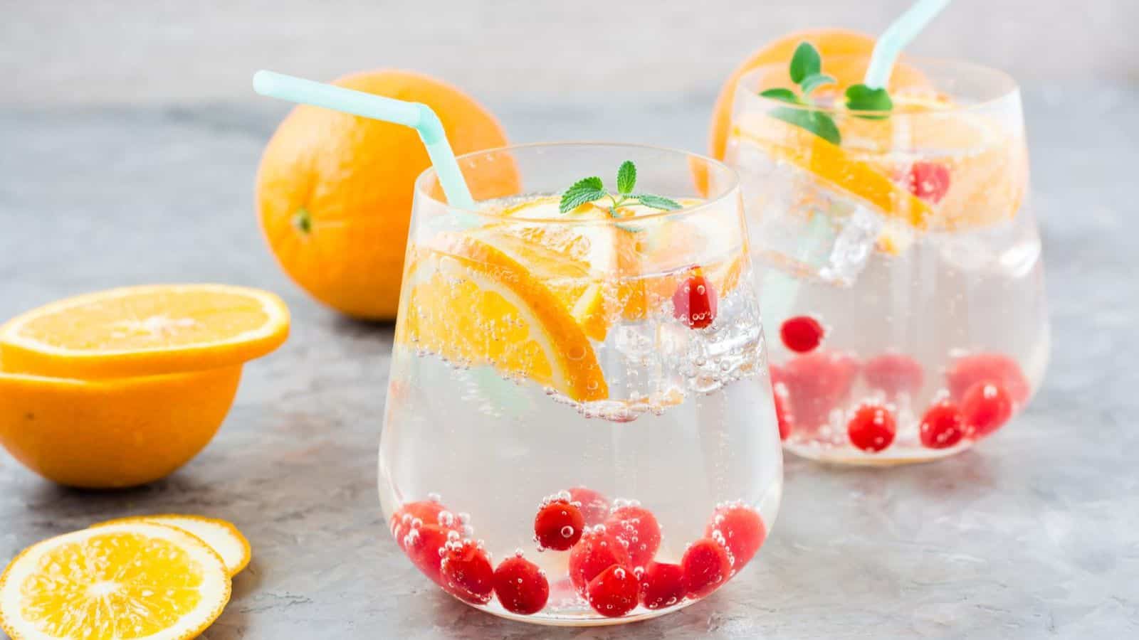 Two clear glasses filled with a fizzy beverage, garnished with orange slices, ice cubes, red berries, and mint leaves. Each glass has a blue straw. Whole and sliced oranges are displayed around the glasses on the gray surface. 