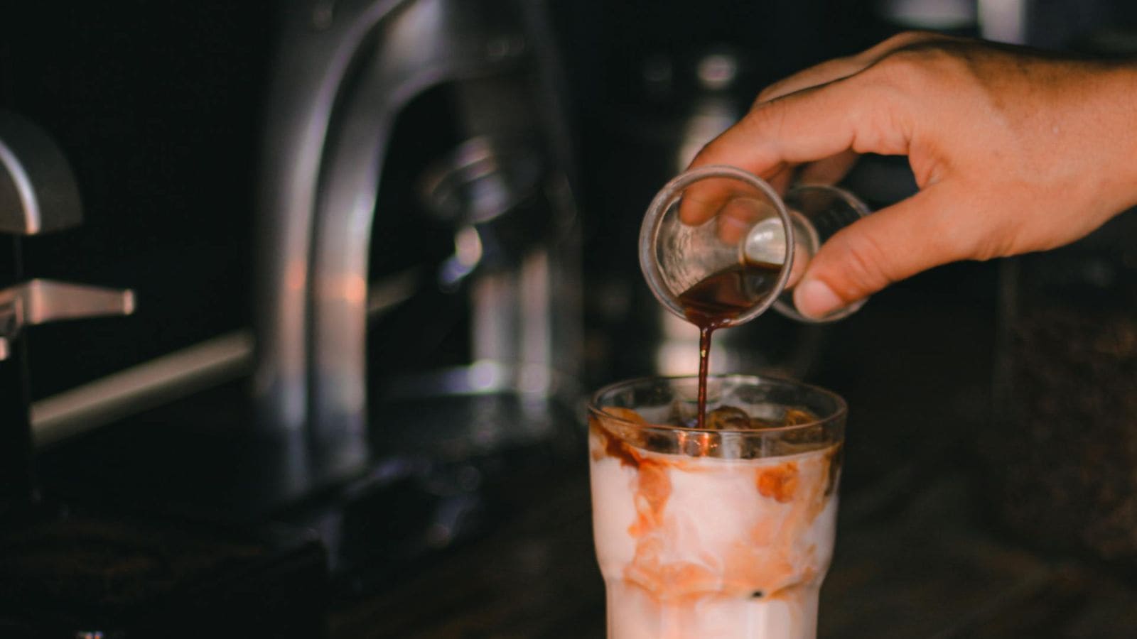 A hand pouring espresso onto a glass of milk