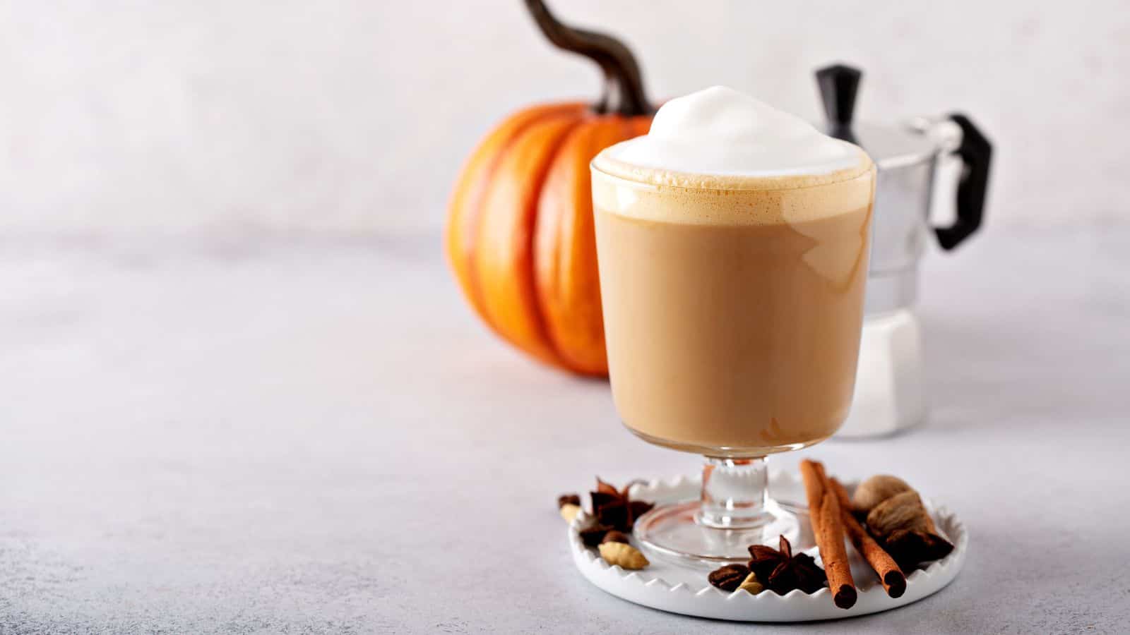 Pumpkin spice latte on a glass placed on top of a coaster with cinnamon and a pumpkin in the background