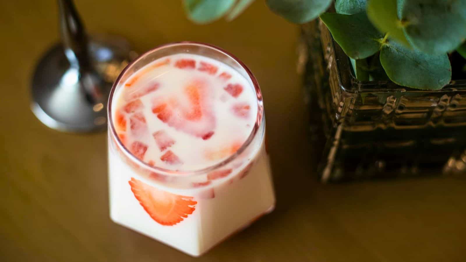 A glass of milky strawberry drink sits on a wooden surface next to a green leafy plant in a black, glass container. The drink contains visible strawberry pieces and is garnished with a strawberry slice on the side of the glass.