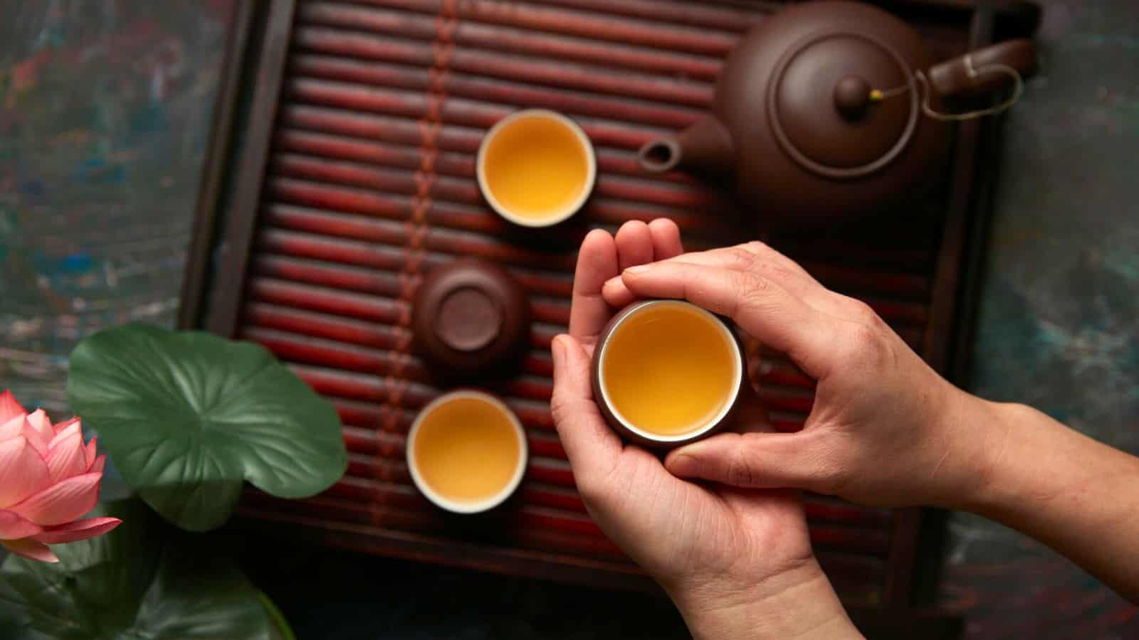 Hands holding a cup of tea over a wooden tray with two additional small cups and a teapot. A green leaf and a pink flower adorn the left side of the tray.