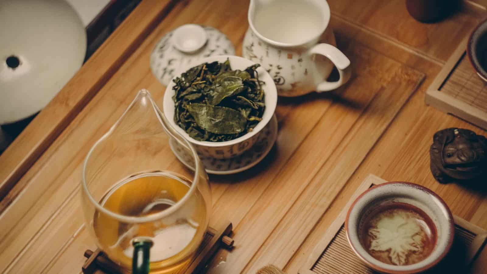 A wooden tea tray holds a glass cup, with dried tea leaves, a small ceramic pitcher with liquid, and a cup with a floral pattern. A ceramic frog ornament and a covered ceramic container are also seen on the tray.