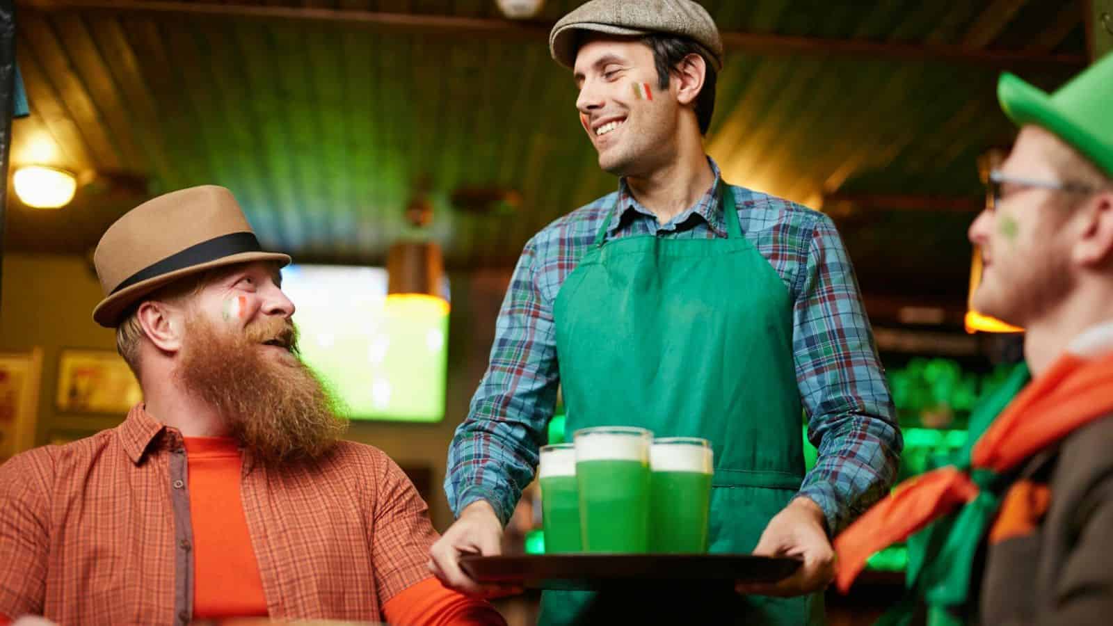 A waiter wearing a green apron serves a tray of fun drinks to two patrons seated at a table in a bar. One patron has a long red beard and hat, while the other wears a green outfit. All have painted cheeks, celebrating in a festive environment.