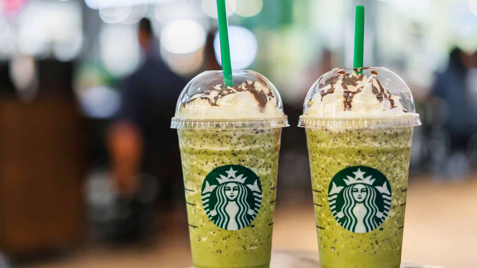 Two green-colored beverages with whipped cream and chocolate drizzle on top, served in clear plastic cups with dome lids and green straws. The logo on the cups indicates they are from Starbucks. The background is blurred.