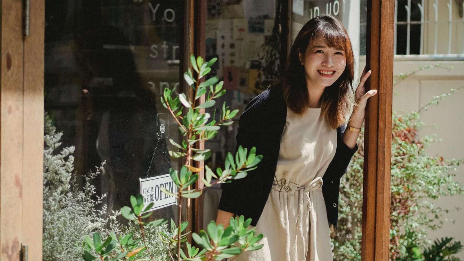 A woman with shoulder-length hair stands at the entrance of a building, slightly leaning against the door frame and smiling. She is wearing a light dress with a black cardigan. A potted plant is in the foreground, and various papers and posters.