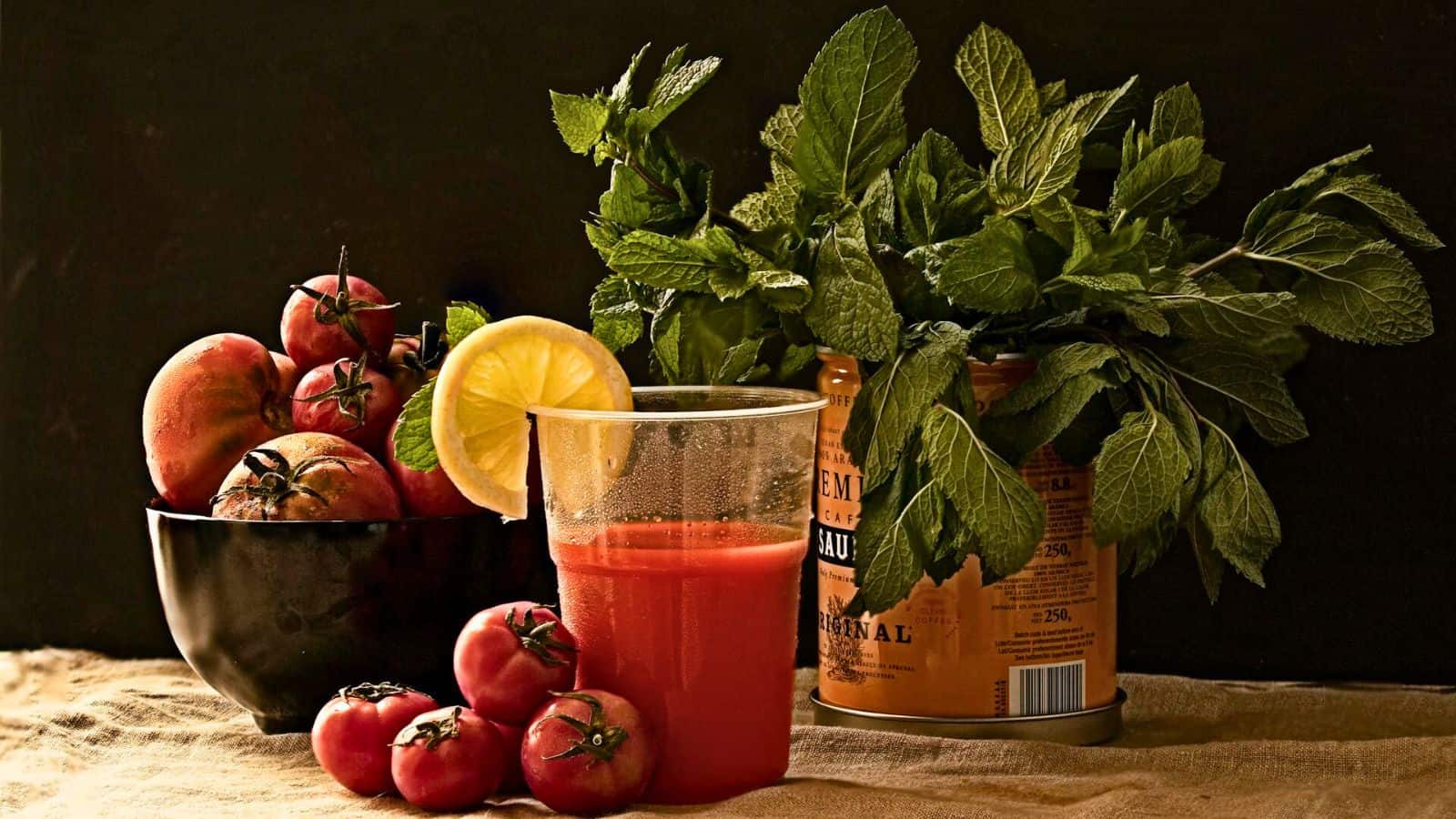 A glass of tomato juice is garnished with a lemon slice and placed on a cloth. Fresh tomatoes are in a black bowl and scattered around. A bunch of green leafy herbs rests in an orange container in the background, all set against a dark backdrop.