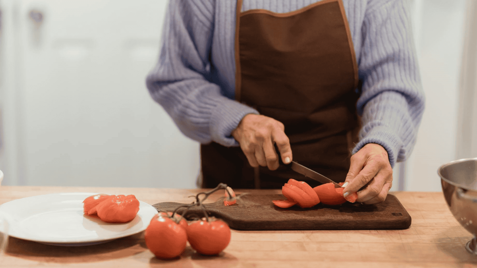 A person wearing a blue sweater and apron is slicing a red bell pepper on a cutting board, pondering unusual plants for winemaking. Several whole bell peppers are nearby on the countertop, alongside a white plate and metal bowl.