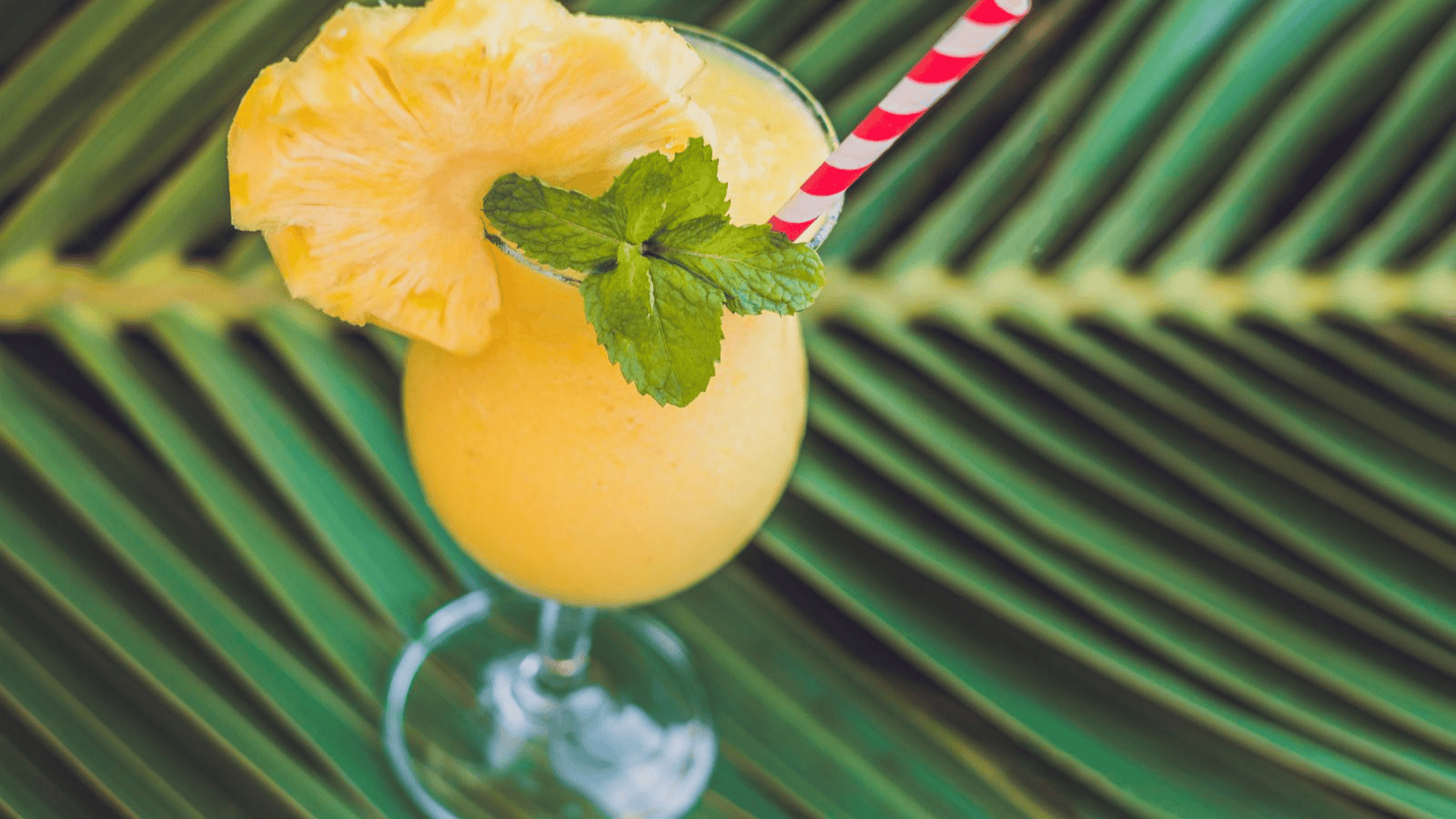 A superfood smoothie served in a glass with a garnish of pineapple slice and mint leaves, elegantly placed on a large green leaf. The vibrant drink includes a red and white striped straw for added charm.