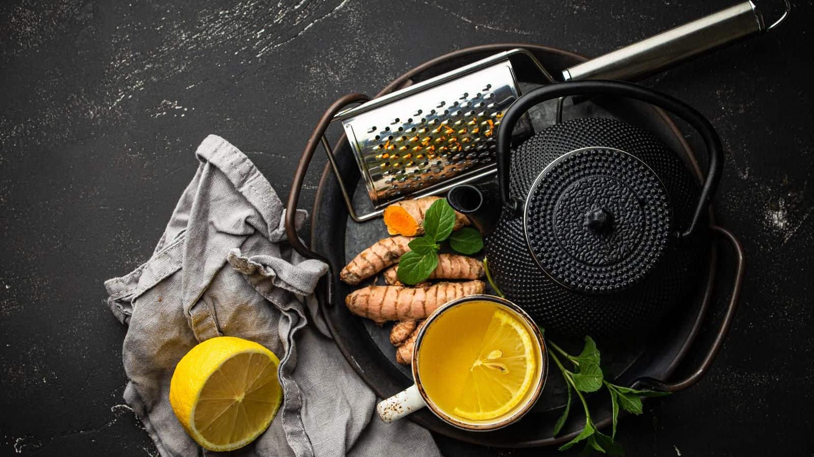 A black teapot, a cup of tea, fresh turmeric roots, a lemon half, a grater, and mint leaves are arranged on a dark surface. A gray cloth napkin is placed adjacent to the lemon.