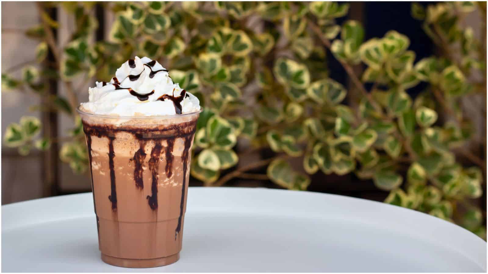 A plastic cup filled with a chocolate milkshake topped with whipped cream and drizzled with chocolate syrup is placed on a white surface. The background includes green and white variegated leaves.