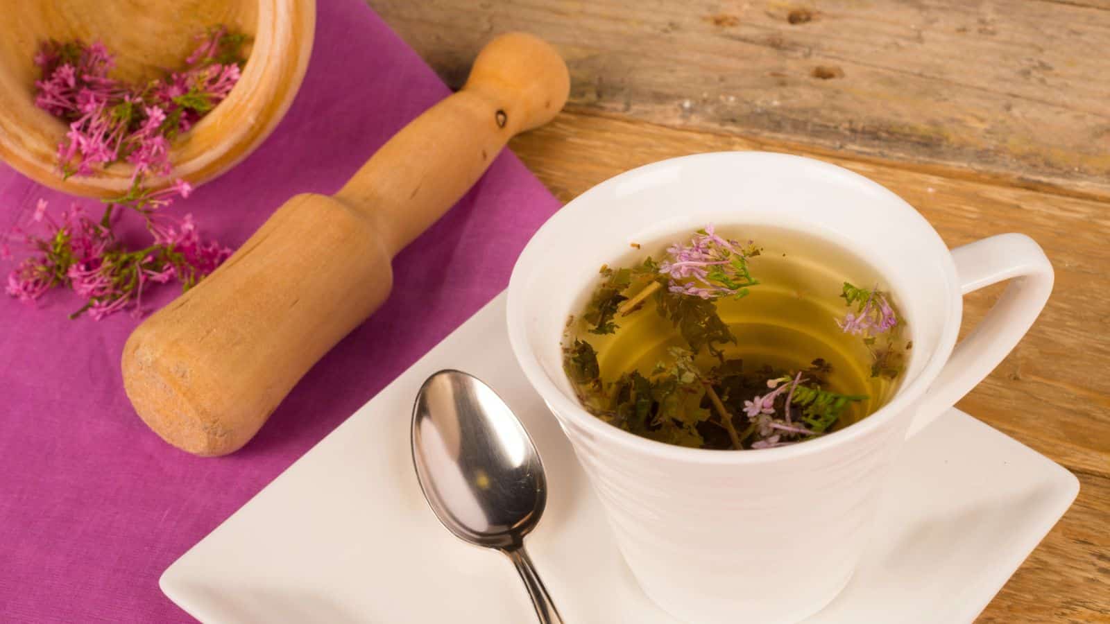 A white cup filled with herbal tea is garnished with small purple flowers and sits on a white saucer with a spoon. To the left, a wooden mortar and pestle rest on a vibrant pink cloth on a wooden surface. Some purple flowers have spilled next to the cup.