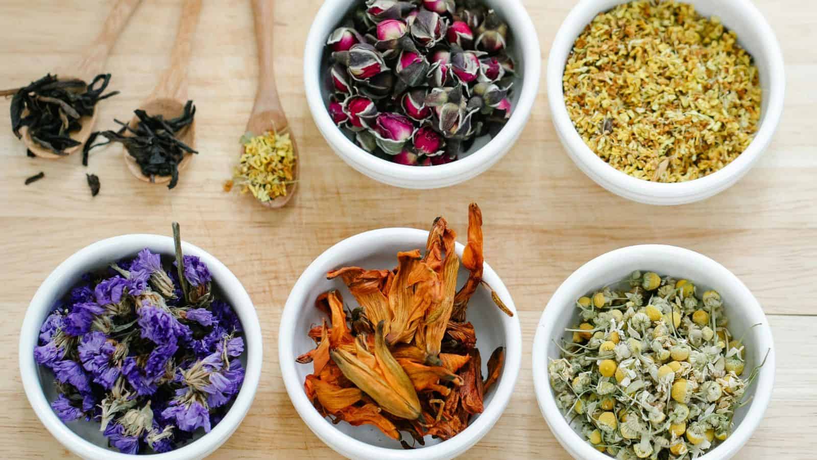 Five small white bowls containing various dried herbs and flowers are arranged on a wooden surface. From top left to bottom right: black tea leaves, rosebuds, yellow flowers, purple flowers, orange petals, and yellow chamomile flowers. Wooden scoops are also visible.