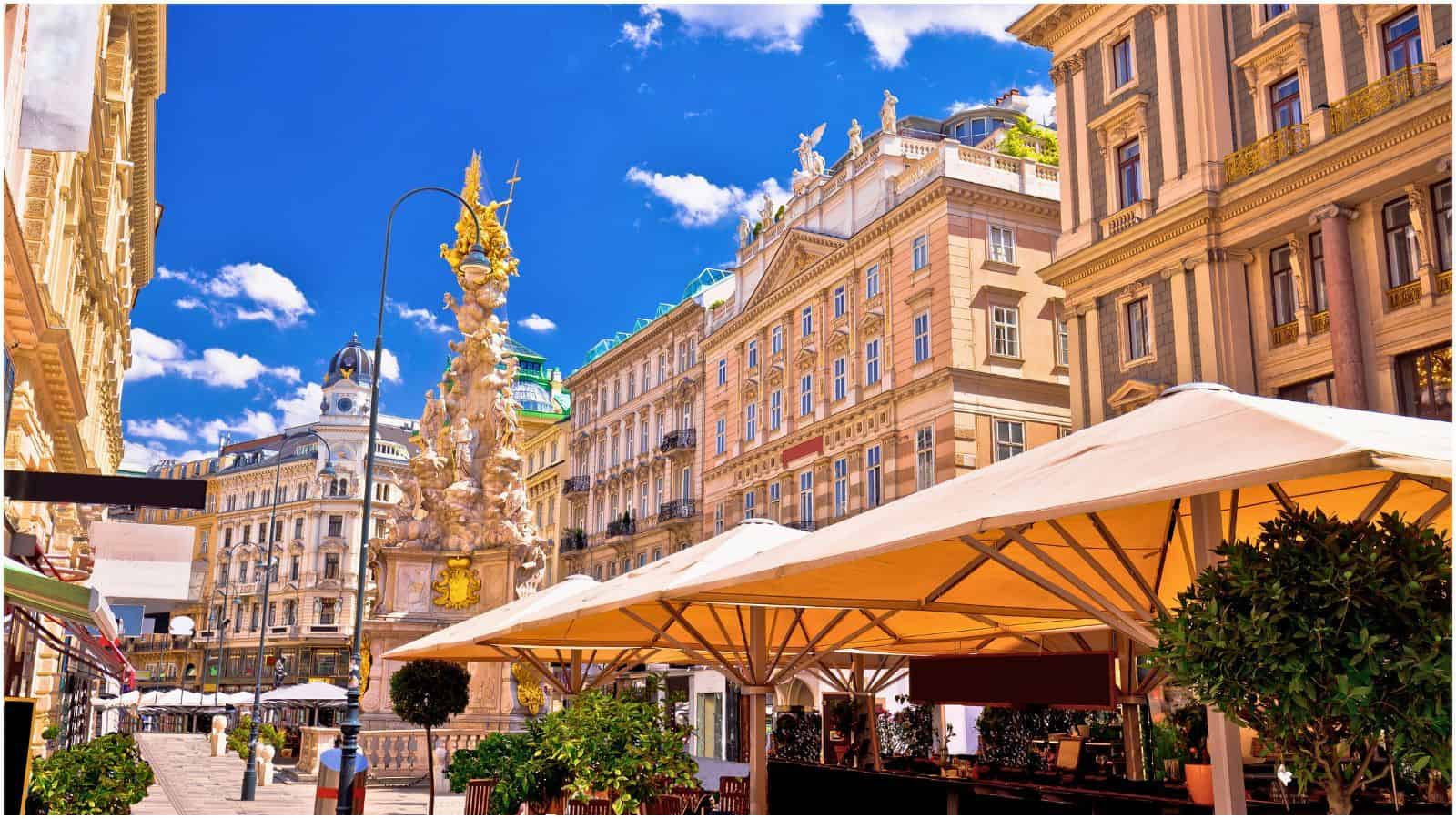 A bustling street scene in Vienna, Austria, with outdoor seating under large umbrellas in the foreground. The street is lined with ornate historic buildings, including a prominent pillar with sculptures. The sky is clear and blue with scattered white clouds.