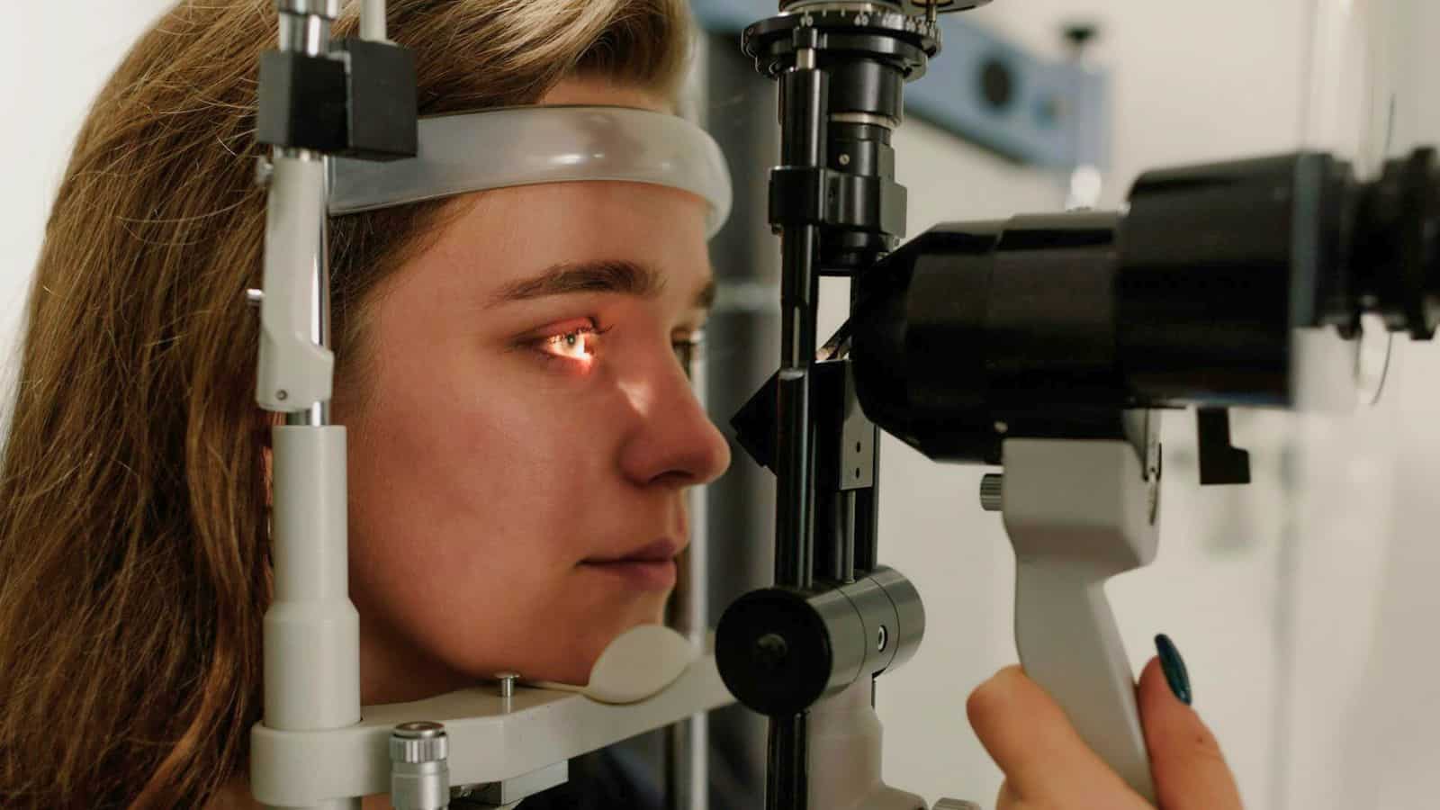 A person with long hair undergoes an eye examination. They are positioned on a chin rest, looking into an ophthalmic device emitting a small beam of light into their eye. The setting is a medical office or clinic.