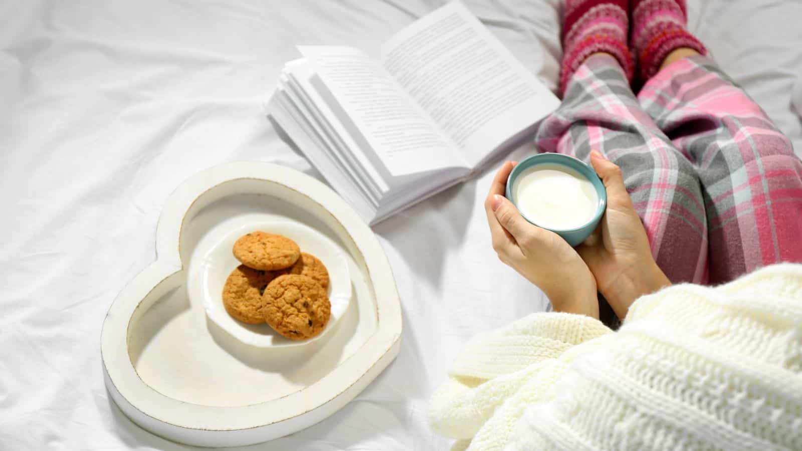 A person wearing plaid pajama pants and cozy socks holds a cup of milk while sitting on a bed with a blanket. Nearby, an open book rests on the bed, and a heart-shaped tray with three cookies is placed next to them.