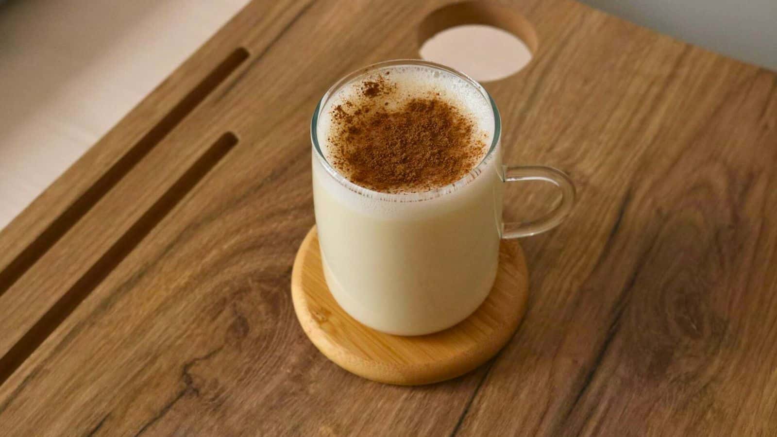 A glass mug filled with milk, topped with a sprinkling of cinnamon, sits on a wooden coaster.