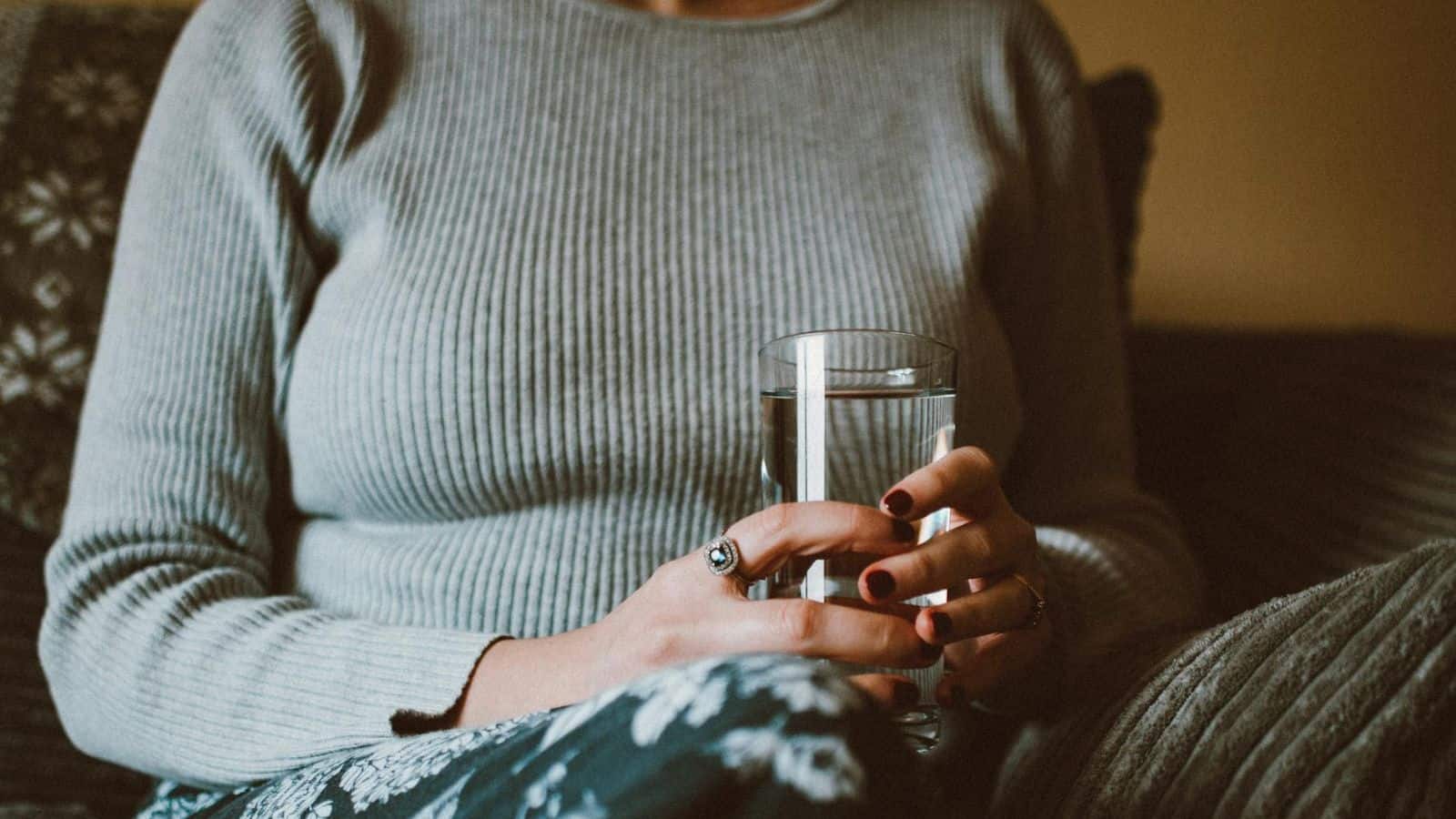 A person wearing a grey ribbed sweater and patterned pants is sitting on a couch, holding a glass of water with both hands. Their fingernails are painted dark red, and they are wearing a ring on their left hand.