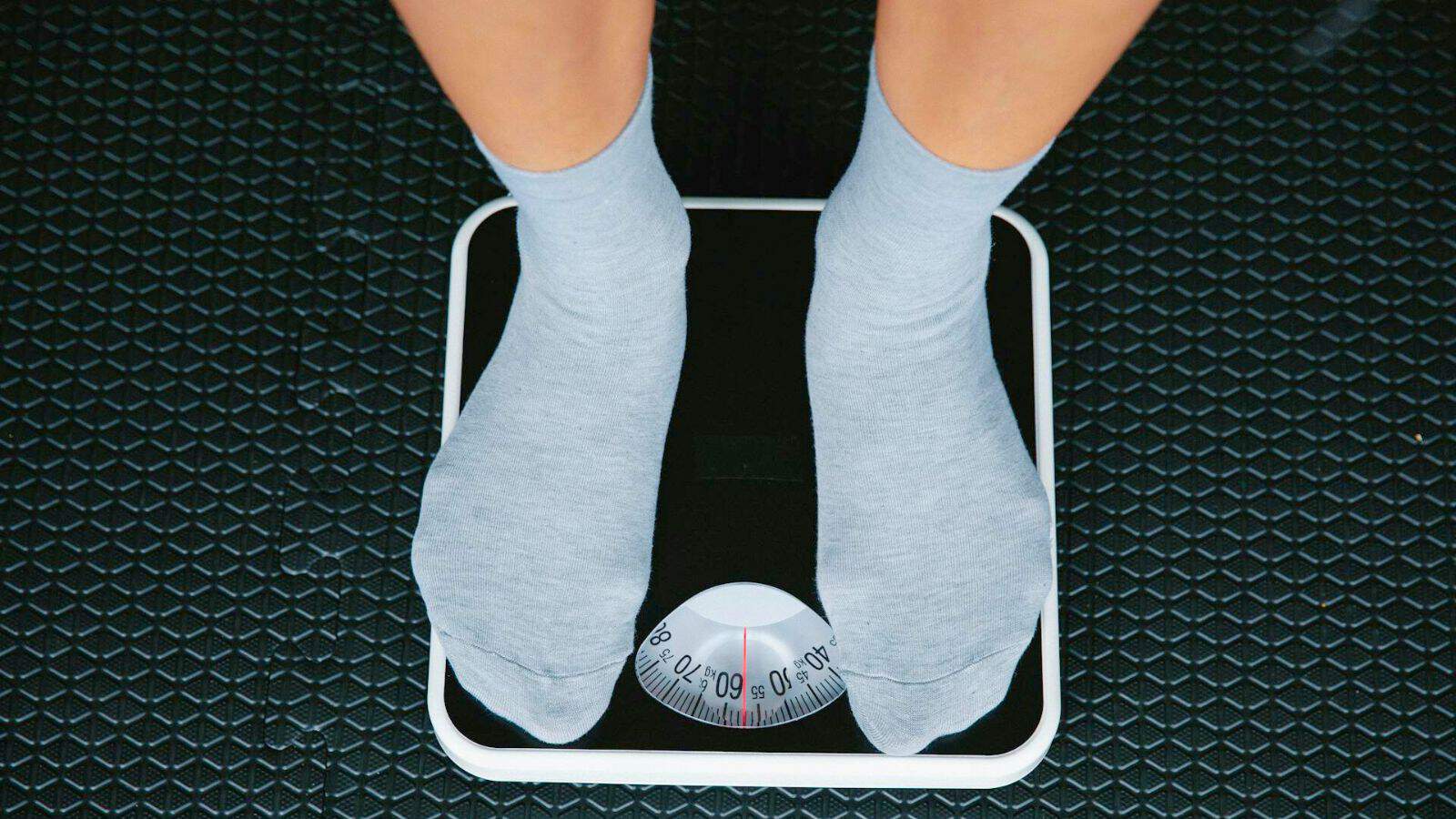Person standing on a black analog weighing scale. Only the feet with grey socks are visible, and the scale reads approximately 60 kilograms. The floor beneath the scale has a textured, dark surface.