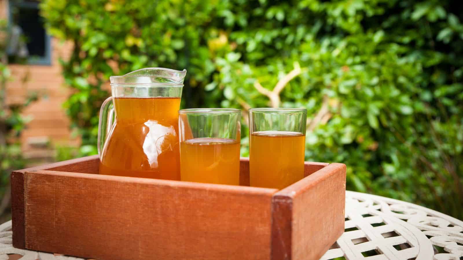 A wooden crate on a white table holds a glass pitcher and two glass cups filled with a kombucha.