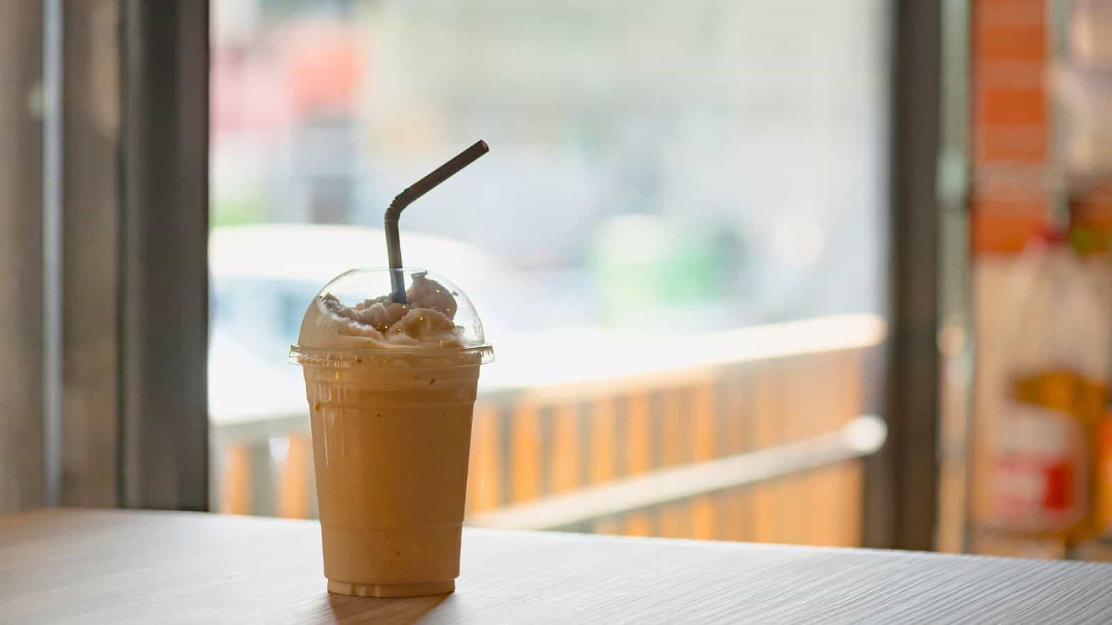 A plastic cup filled with a light brown iced beverage, topped with whipped cream, sits on a wooden table. The cup has a domed lid with a bent black straw inserted. The background features a blurred view of a window and an outdoor scene.